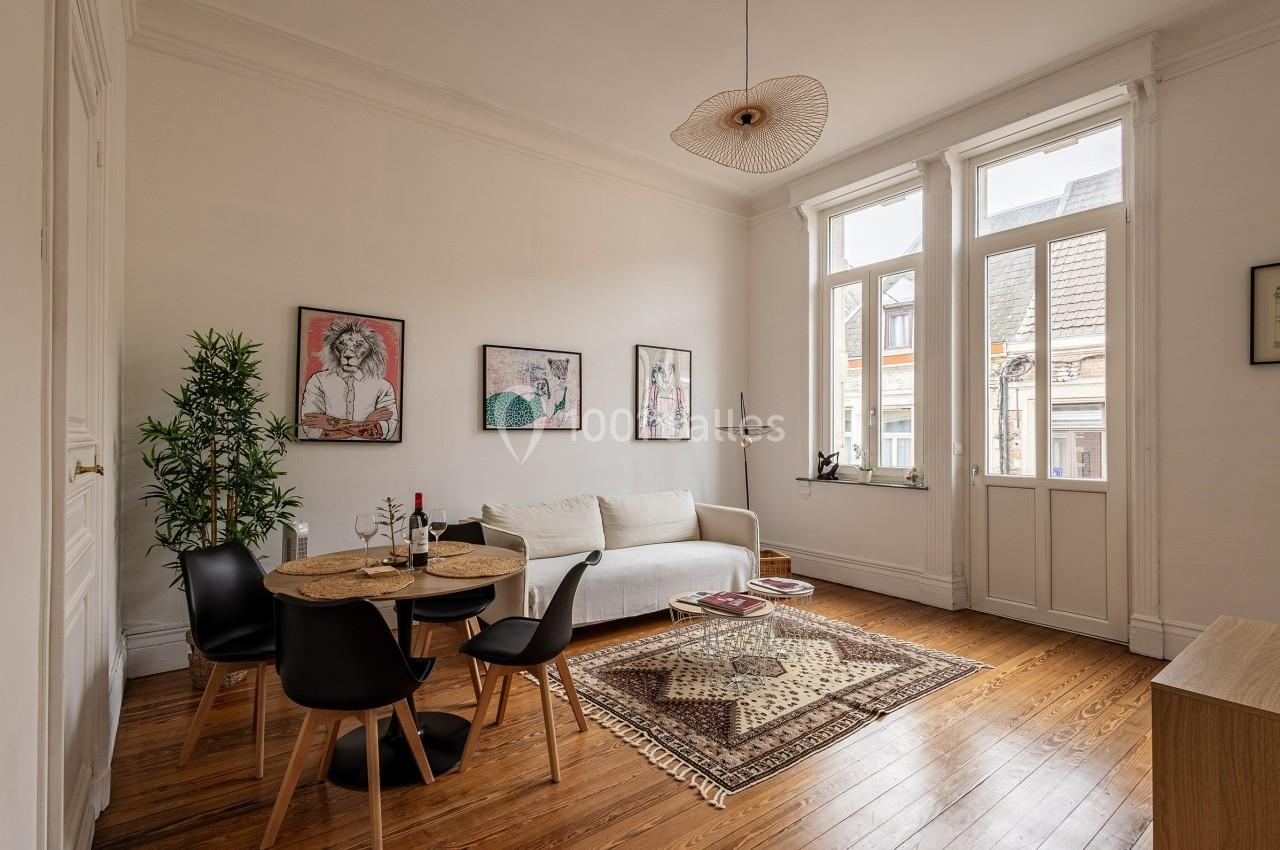 Salon lumineux avec parquet, canapé blanc, table ronde avec chaises noires, décorations murales et grandes fenêtres.