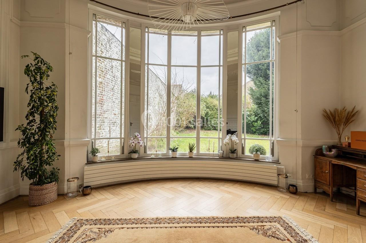 Grande baie vitrée arrondie donnant sur un jardin, entourée de plantes et décorée d'un tapis et de meubles en bois.