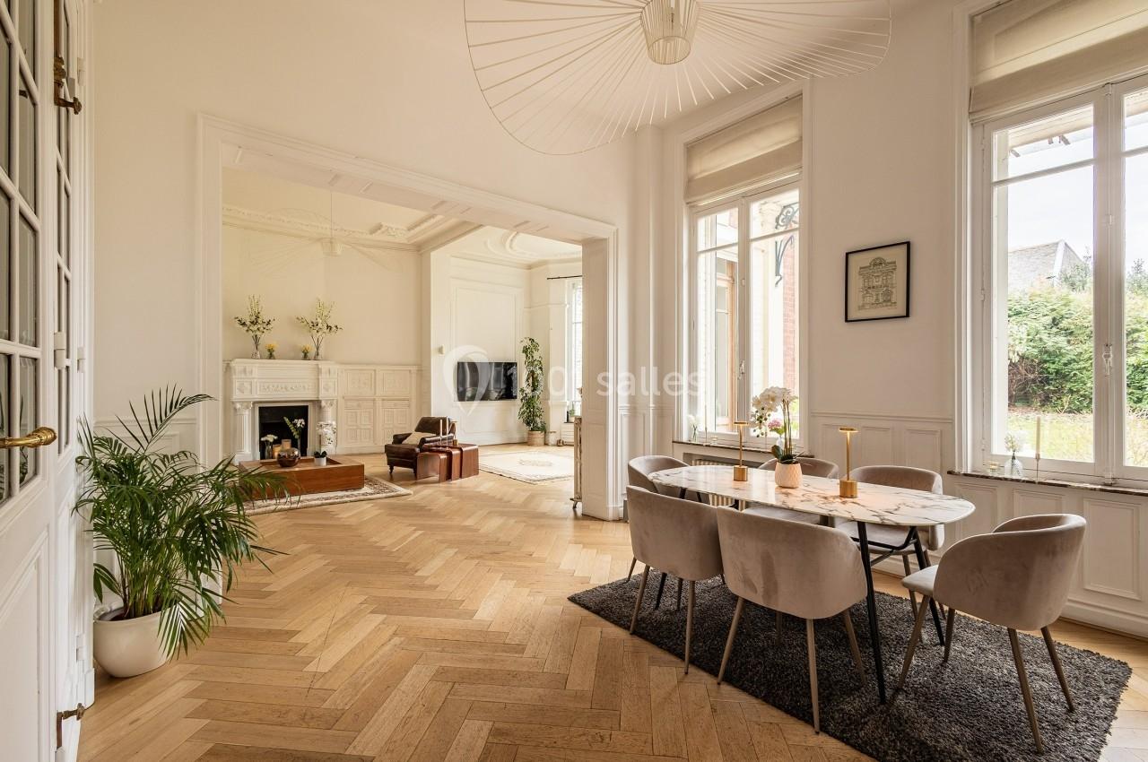 Salle à manger lumineuse avec table, chaises, parquet en bois clair, cheminée et grandes fenêtres donnant sur un jardin.