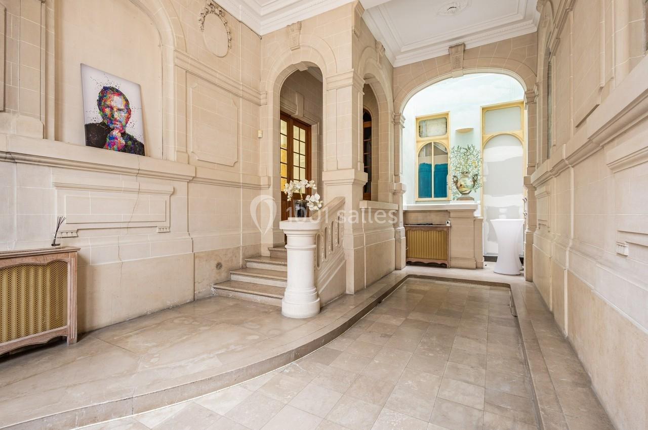 Entrée lumineuse avec murs en pierre beige, escalier, tableau coloré et vue sur une cour intérieure.