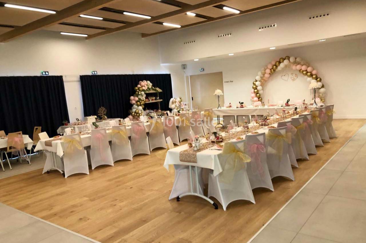 Salle décorée pour une réception avec tables alignées, nappes blanches, chaises ornées de nœuds et arche de ballons.
