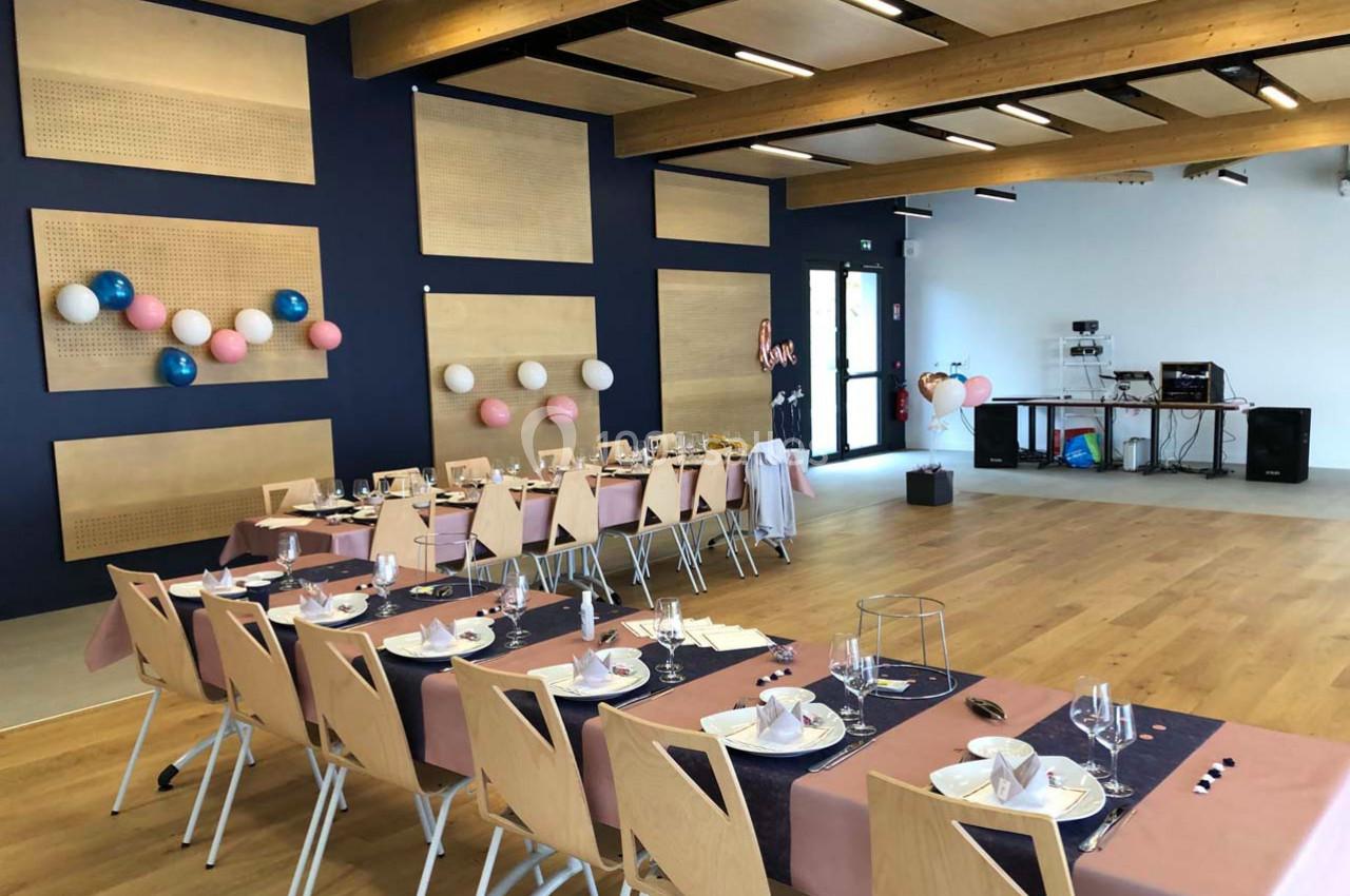 Salle décorée pour un événement, avec tables dressées, nappes roses et bleues, et ballons accrochés aux murs.