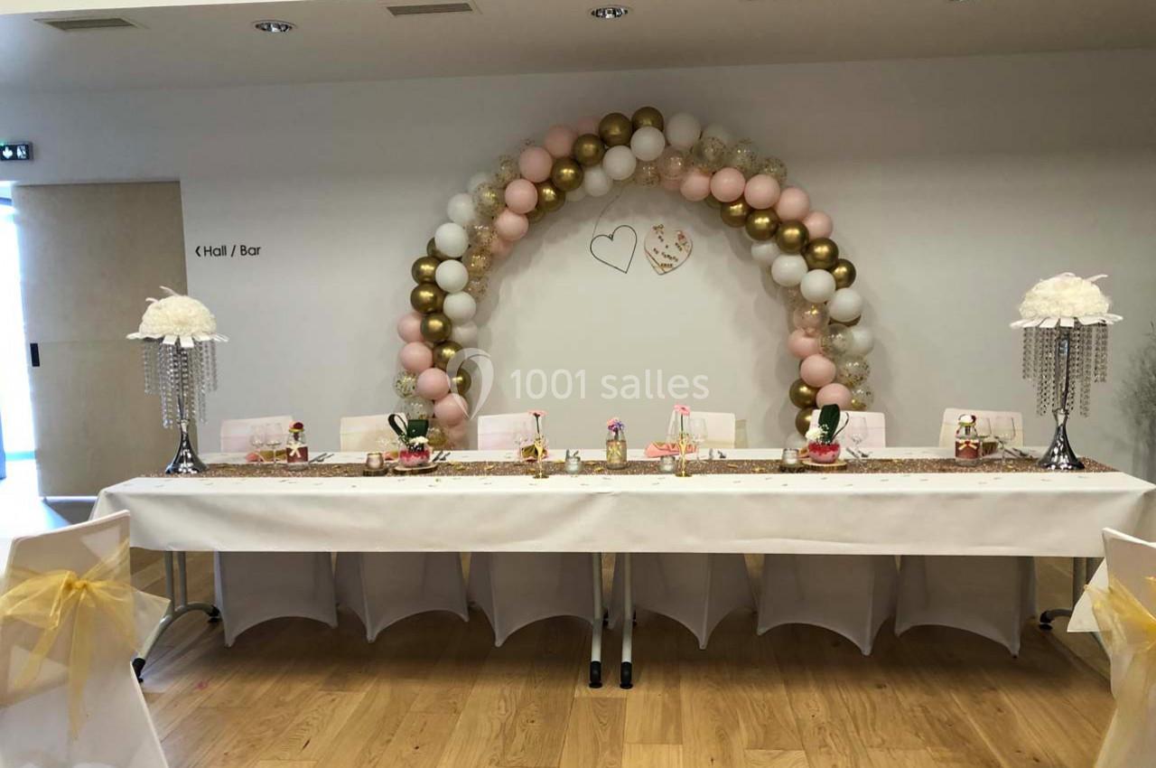 Table décorée pour un événement avec arche de ballons roses et dorés en arrière-plan, et centres de table élégants.