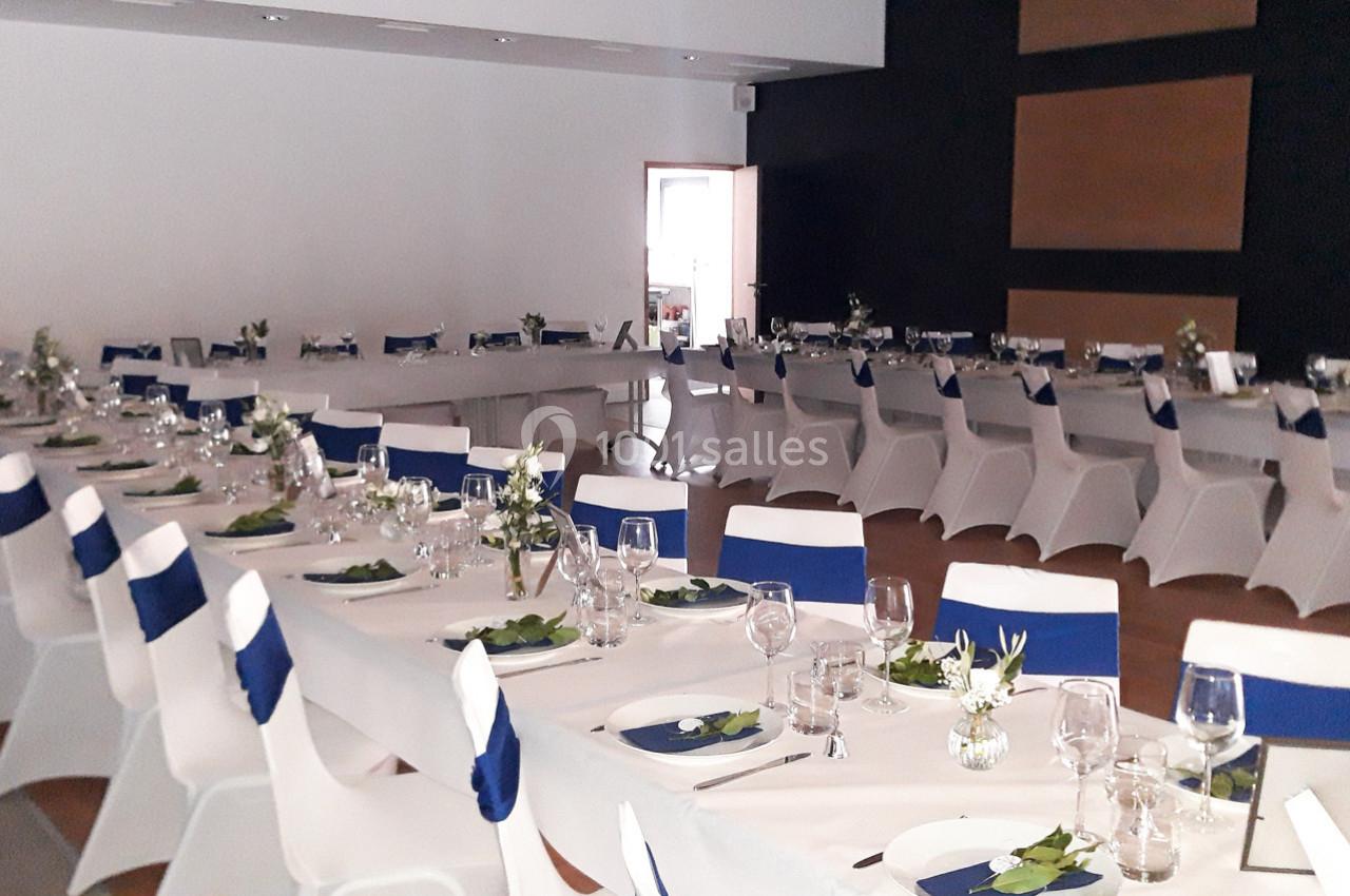 Salle de réception décorée avec des tables dressées, nappes blanches, serviettes bleues et arrangements floraux.