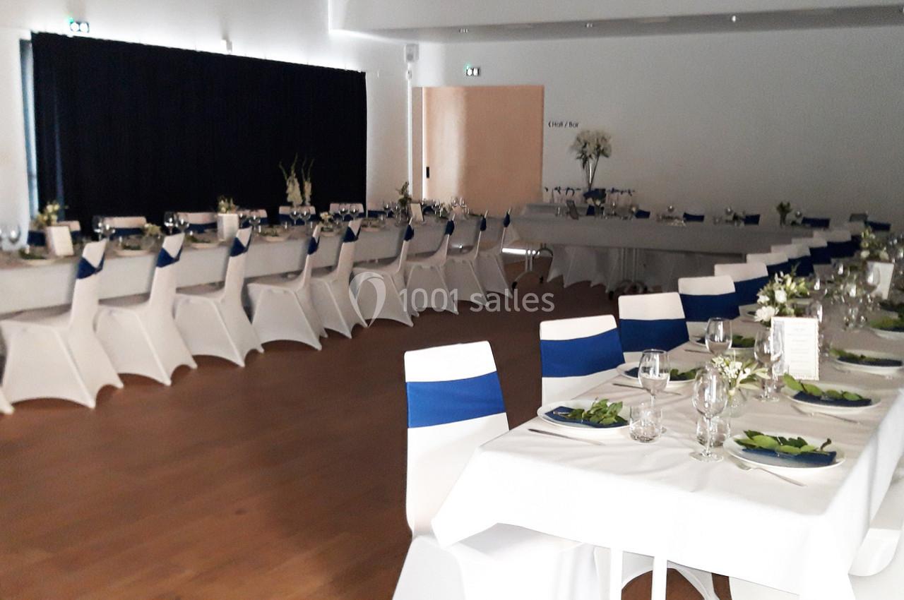Salle de réception avec tables dressées, nappes blanches, chaises décorées de rubans bleus et arrangements floraux.