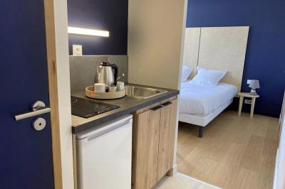 Kitchenette équipée avec évier, plaques de cuisson, bouilloire et vue sur une chambre avec lit double et parquet.