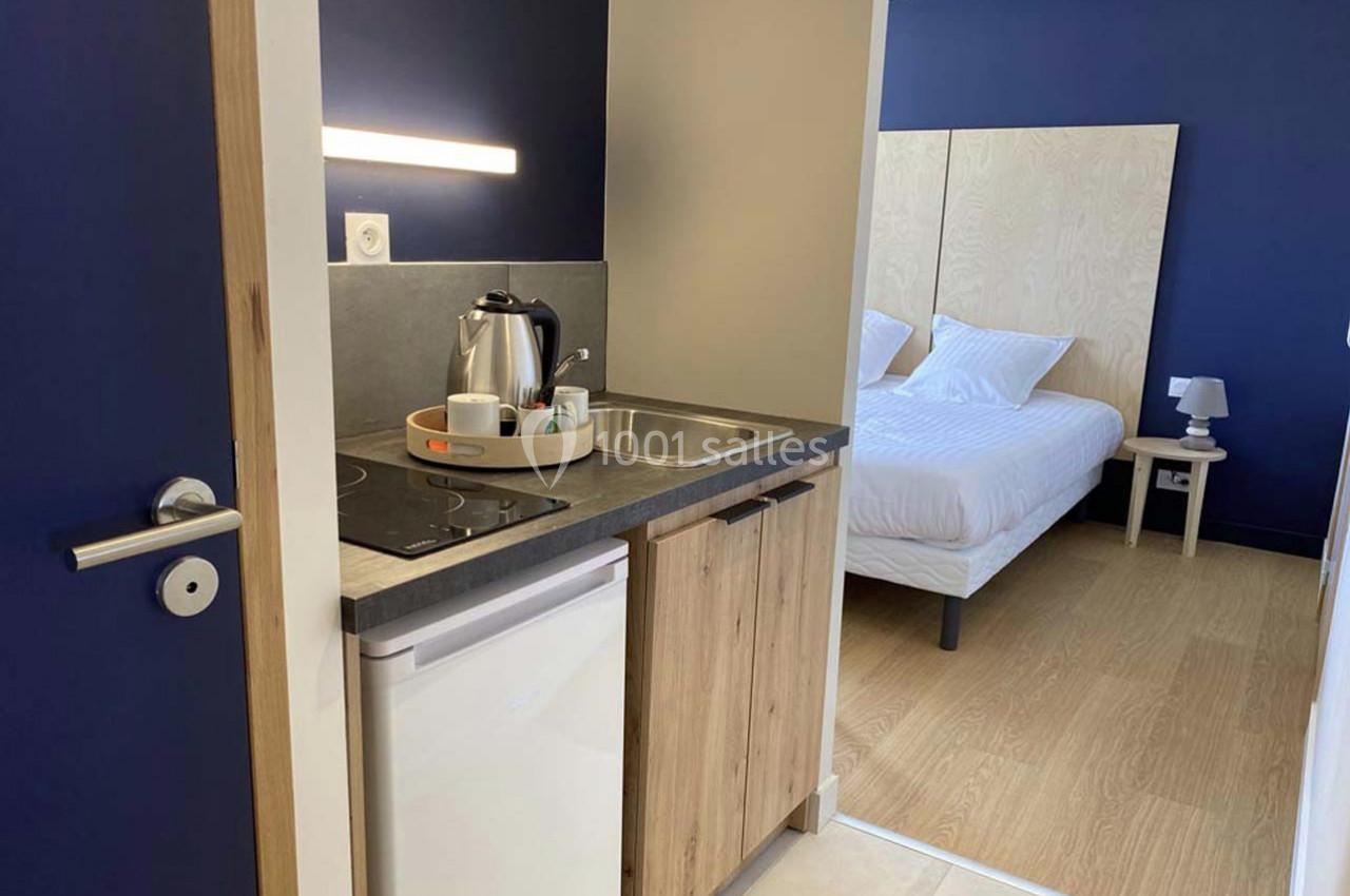 Kitchenette équipée avec évier, plaques de cuisson, bouilloire et vue sur une chambre avec lit double et parquet.