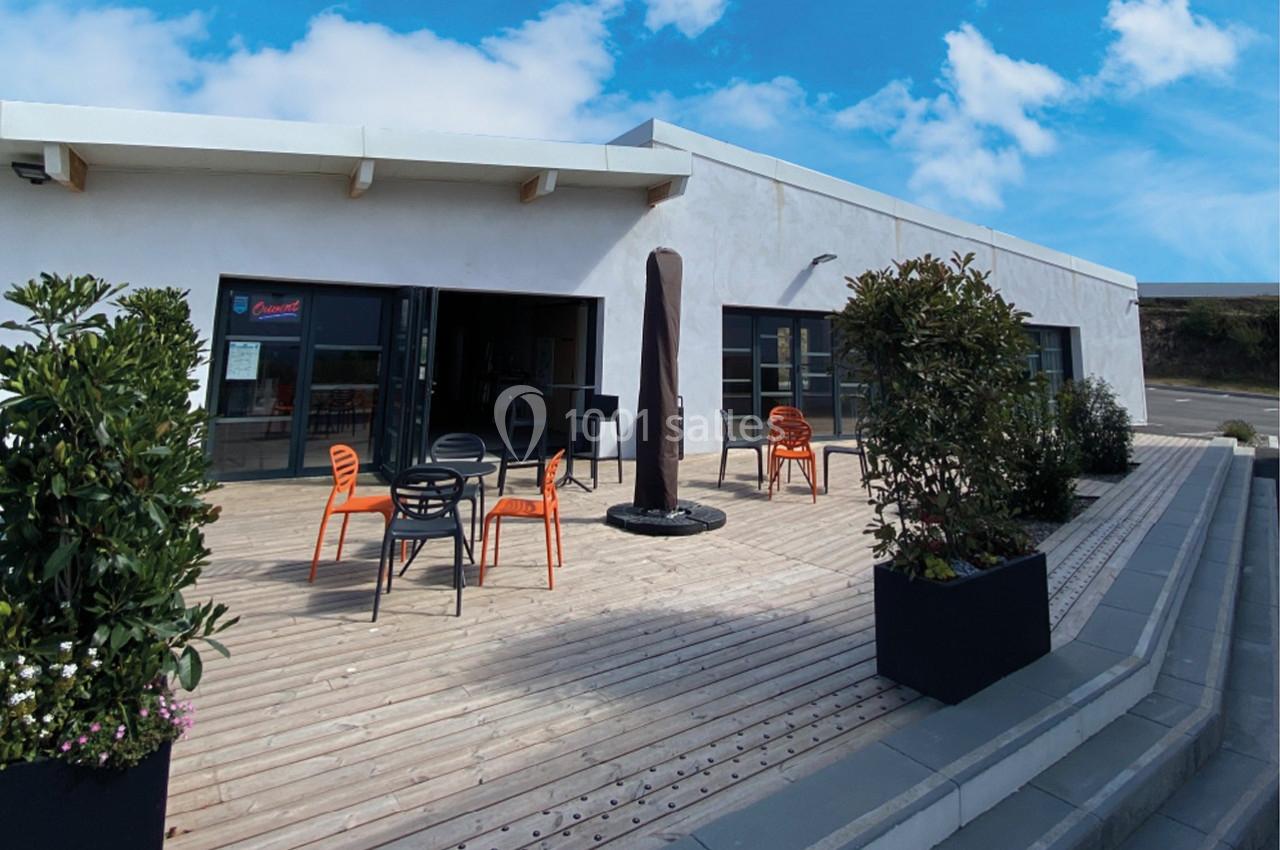 Terrasse en bois avec tables et chaises orange, entourée de plantes, devant un bâtiment moderne sous un ciel bleu.