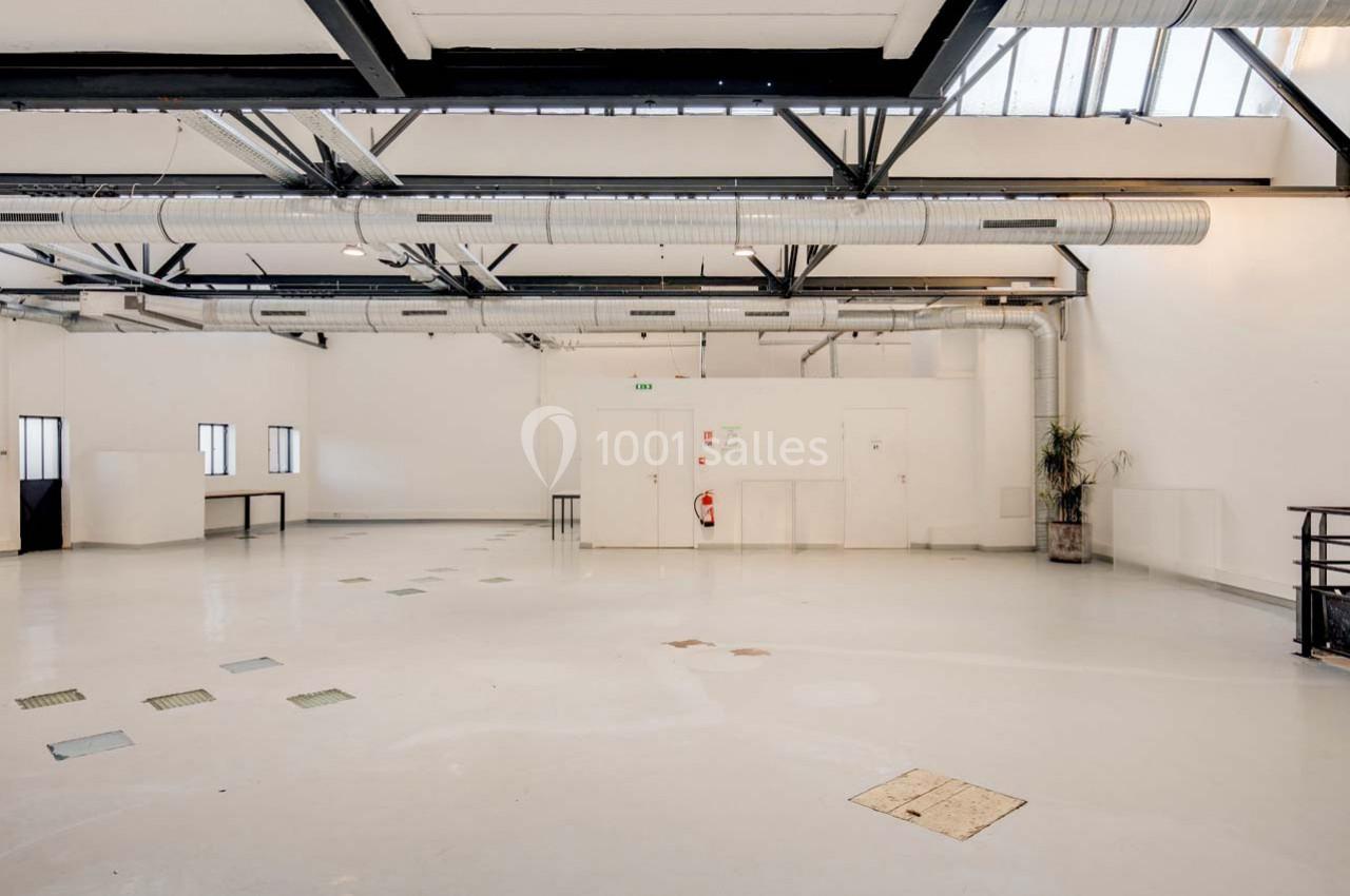 Intérieur d'un grand espace industriel lumineux avec plafond en poutres métalliques, conduits apparents et murs blancs.