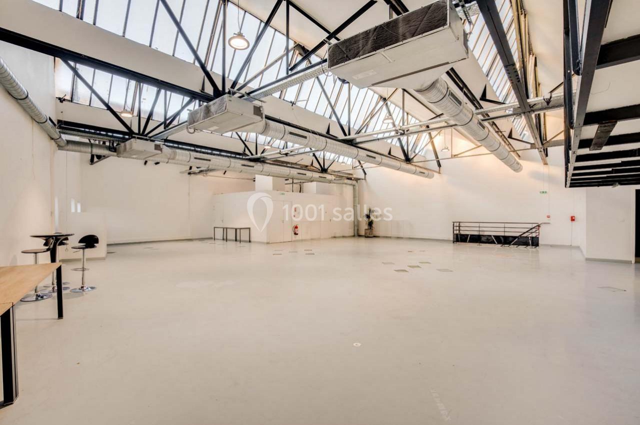 Intérieur d'un grand espace industriel lumineux avec plafond en verrière, poutres métalliques et sol en béton clair.