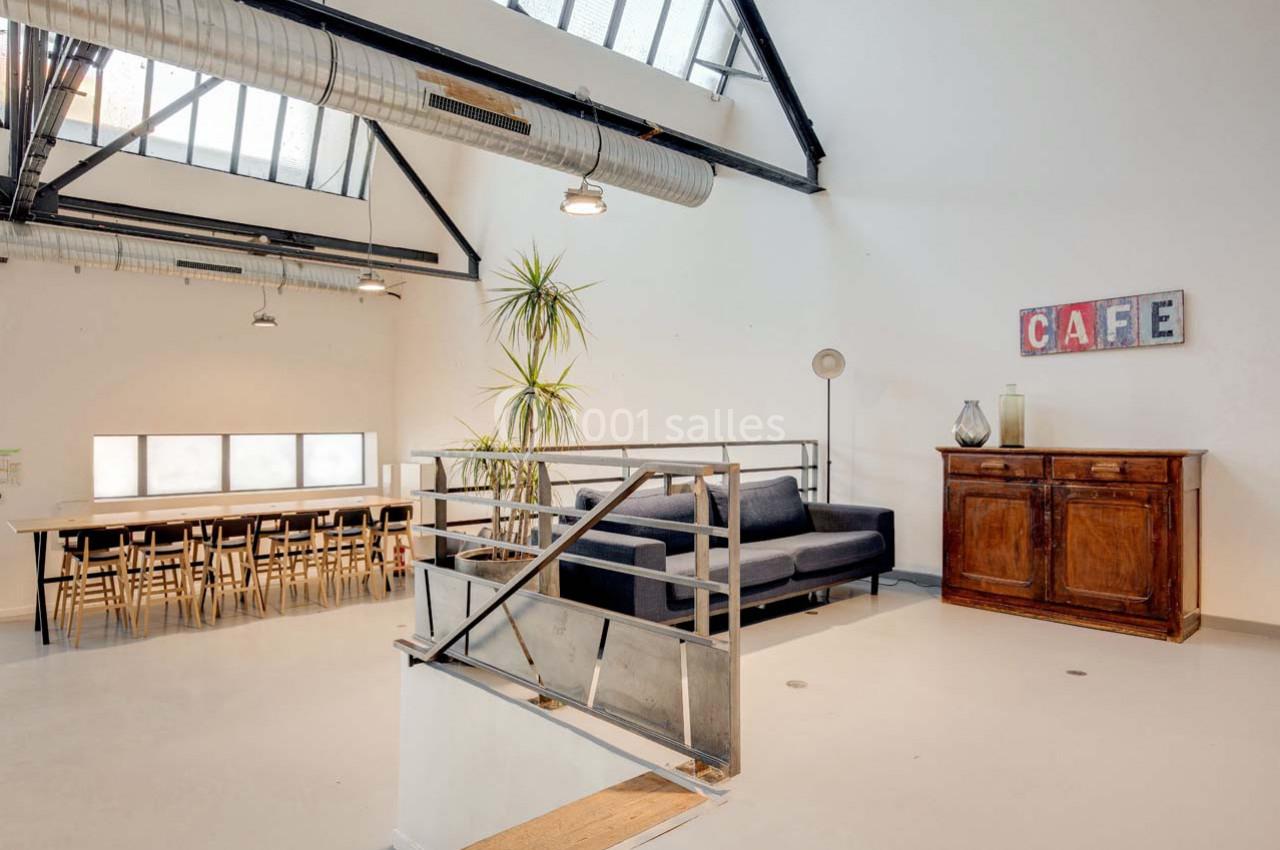 Espace lumineux avec grande table en bois, canapé gris, meuble ancien et plantes sous une verrière industrielle.