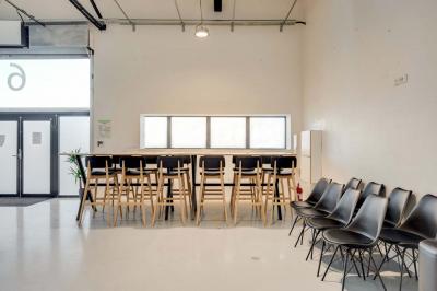 Salle lumineuse avec rangées de chaises, tables hautes et buffet en arrière-plan, sous un plafond industriel.