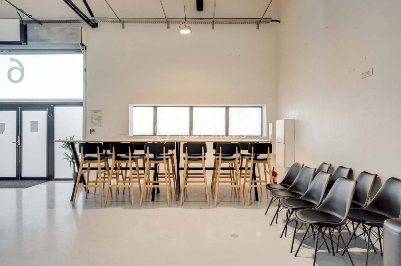 Salle lumineuse avec un comptoir haut entouré de tabourets en bois et une rangée de chaises noires alignées contre le mur.