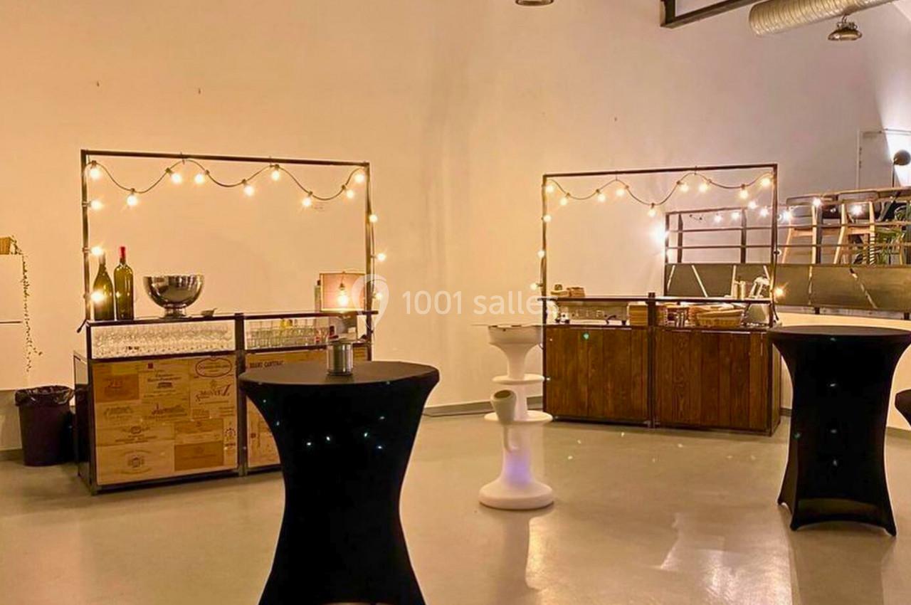 Salle décorée avec des guirlandes lumineuses, tables hautes noires et comptoirs en bois pour un événement.