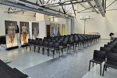 Salle lumineuse avec rangées de chaises, tables hautes et buffet en arrière-plan, sous un plafond industriel.
