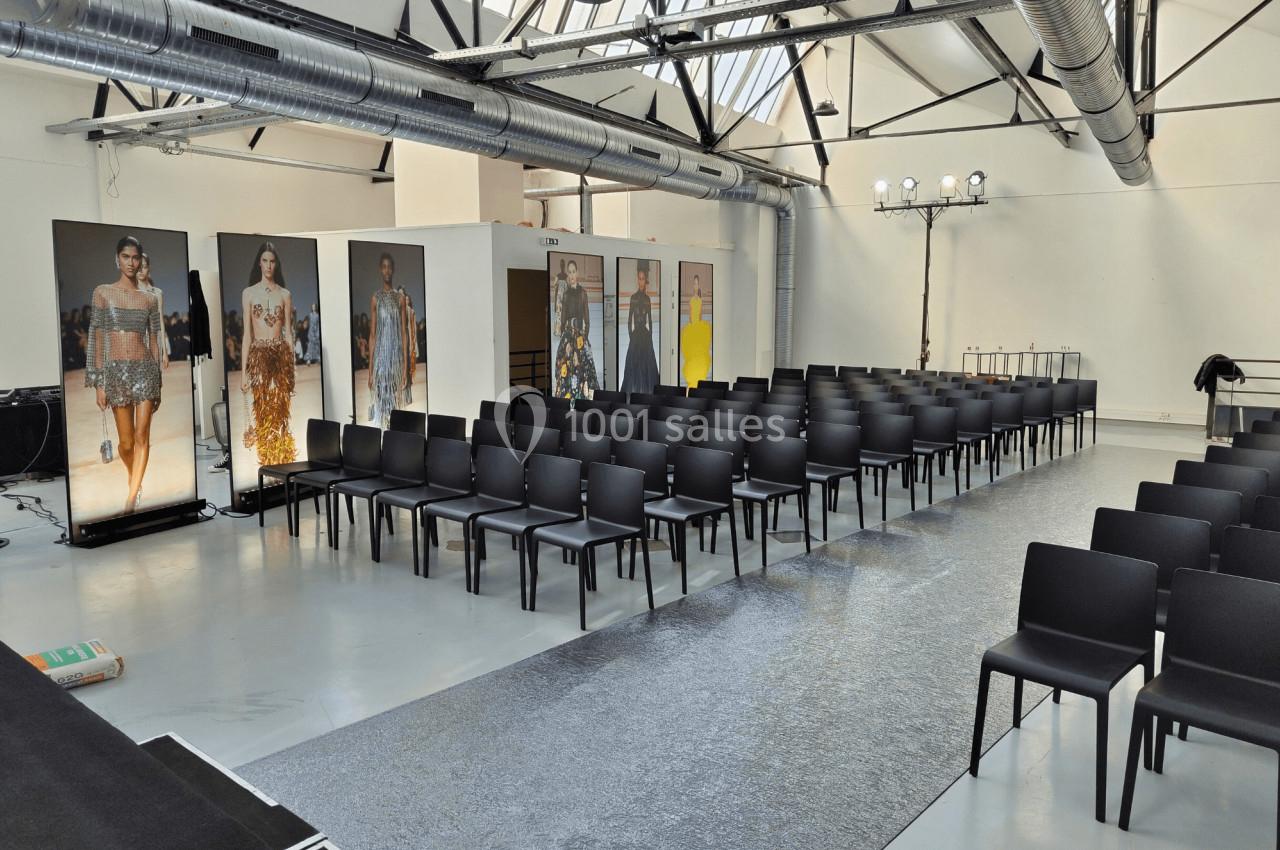 Salle lumineuse aménagée pour un défilé de mode avec rangées de chaises noires et affiches de mannequins en arrière-plan.