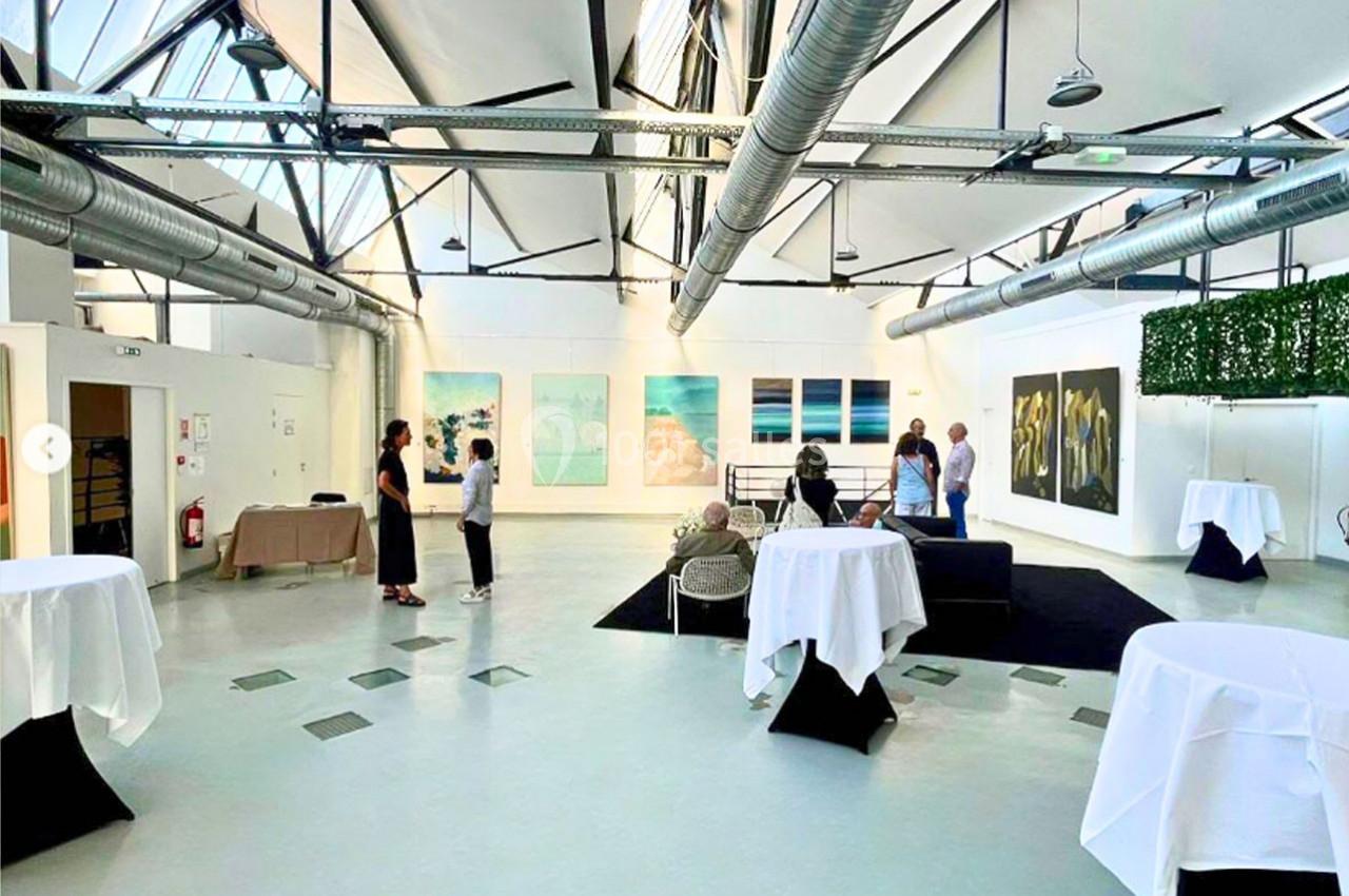 Salle lumineuse avec des œuvres d'art accrochées aux murs, des tables hautes et quelques personnes discutant.