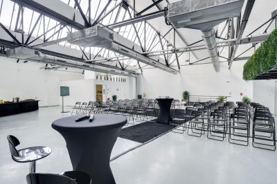 Salle lumineuse avec rangées de chaises, tables hautes et buffet en arrière-plan, sous un plafond industriel.
