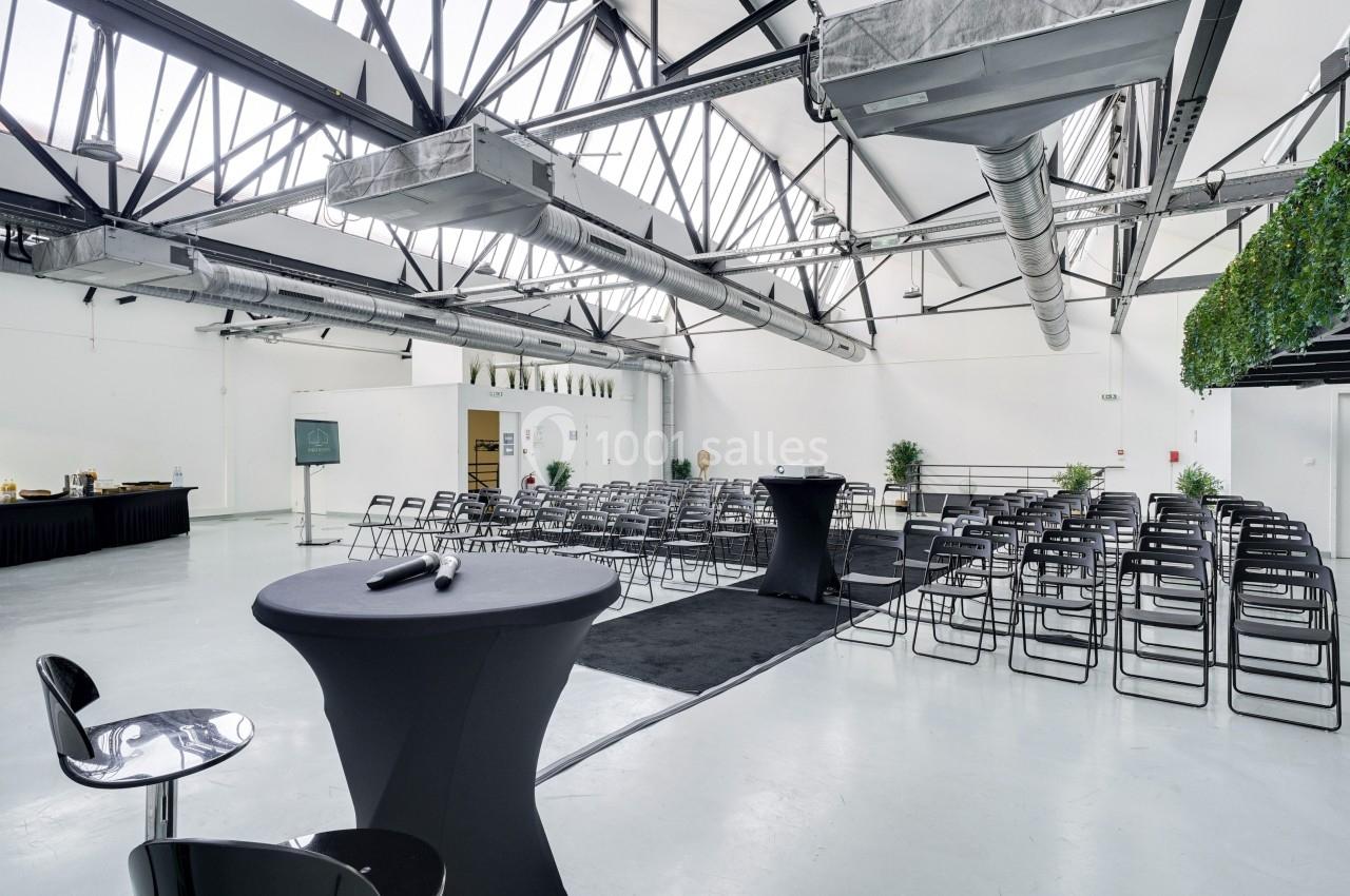 Salle lumineuse avec rangées de chaises, tables hautes et buffet en arrière-plan, sous un plafond industriel.
