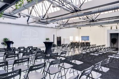 Salle lumineuse avec rangées de chaises, tables hautes et buffet en arrière-plan, sous un plafond industriel.
