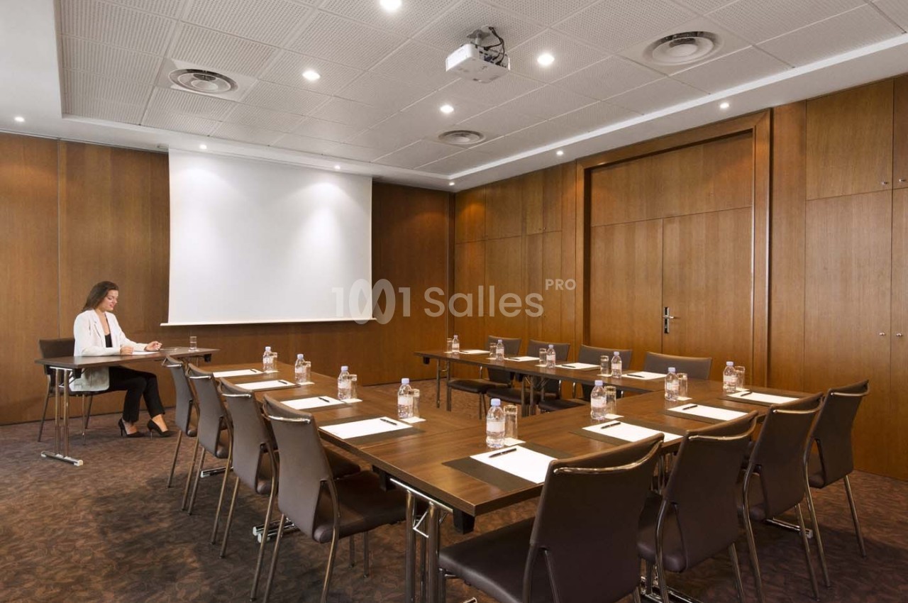 Salle de réunion équipée avec tables, chaises, bouteilles d'eau et écran de projection, une personne assise seule.