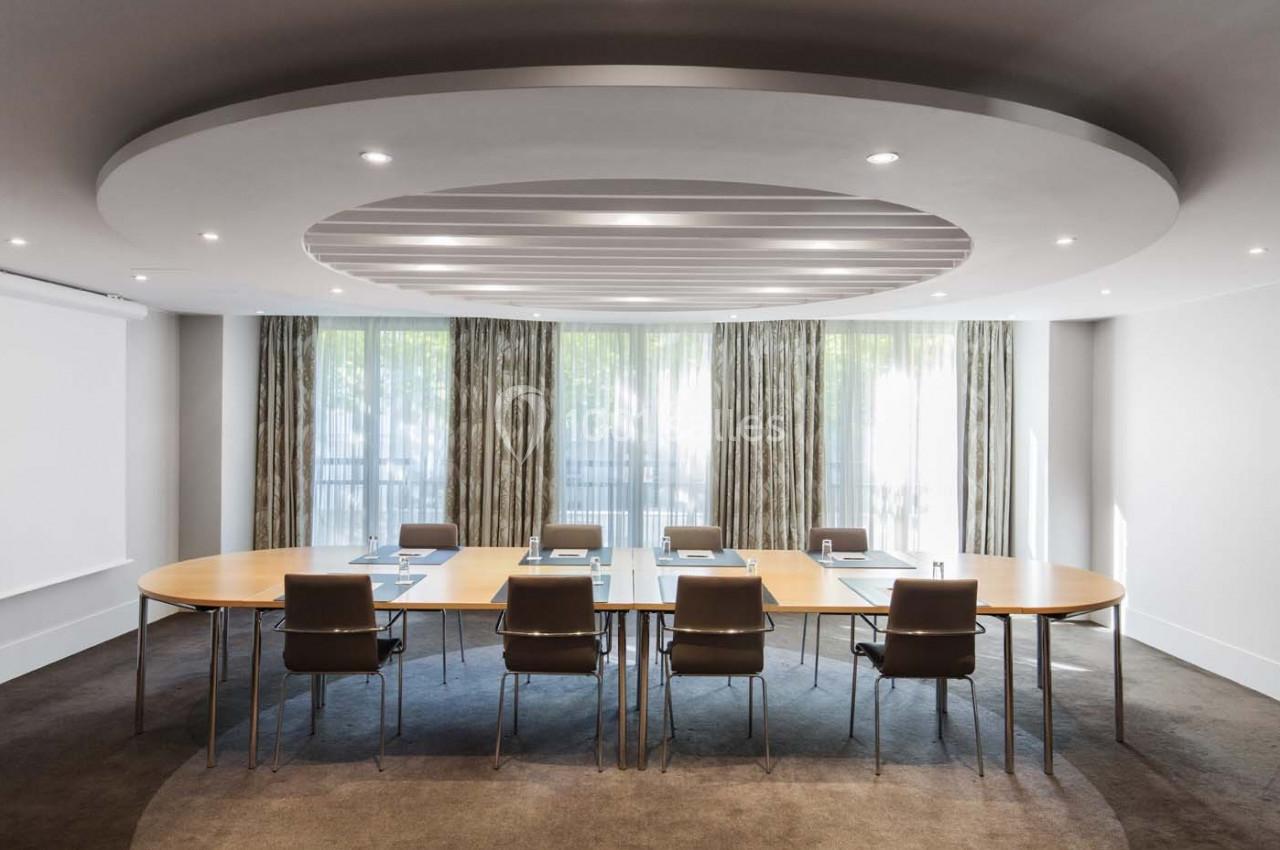 Salle de réunion lumineuse avec une grande table ovale, chaises en cuir, rideaux et éclairage encastré au plafond.