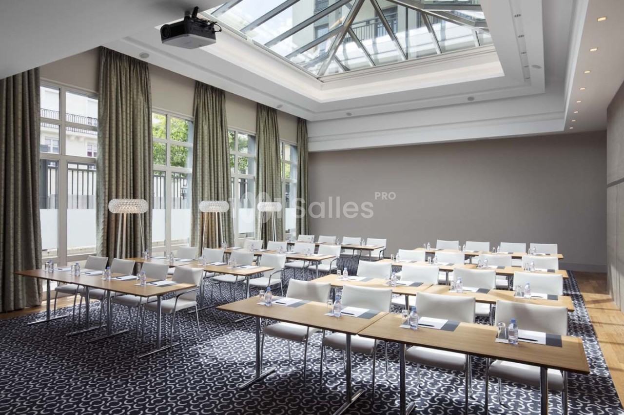 Salle de conférence lumineuse avec des tables alignées, des chaises blanches et un plafond vitré.