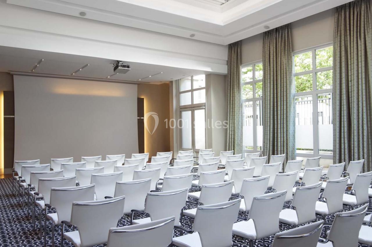 Salle de conférence lumineuse avec des chaises blanches alignées, un écran de projection et une verrière au plafond.
