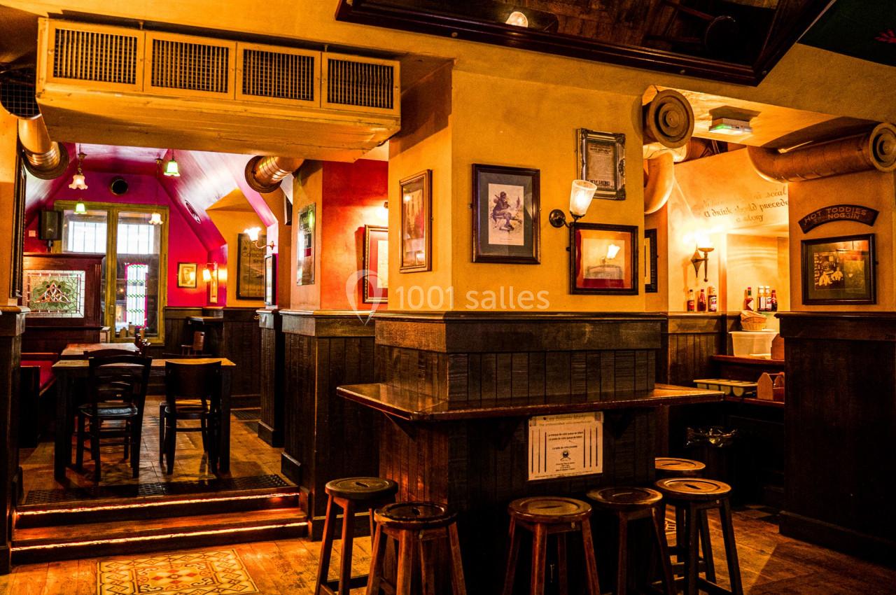 Intérieur d'un bar chaleureux avec des tabourets en bois, des tables, et des murs décorés de cadres et d'affiches.