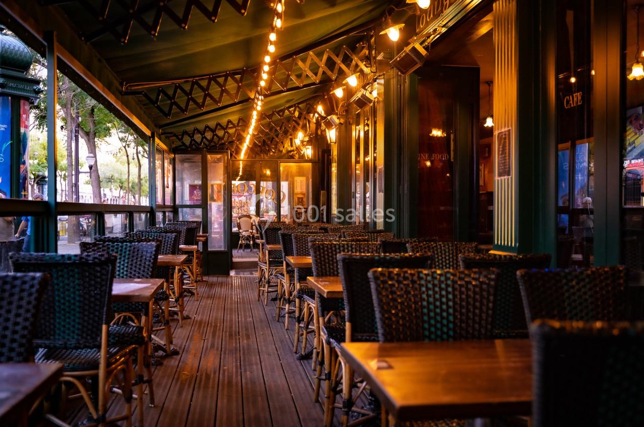 Terrasse de café vide avec tables et chaises en osier, éclairée par des guirlandes lumineuses au crépuscule.