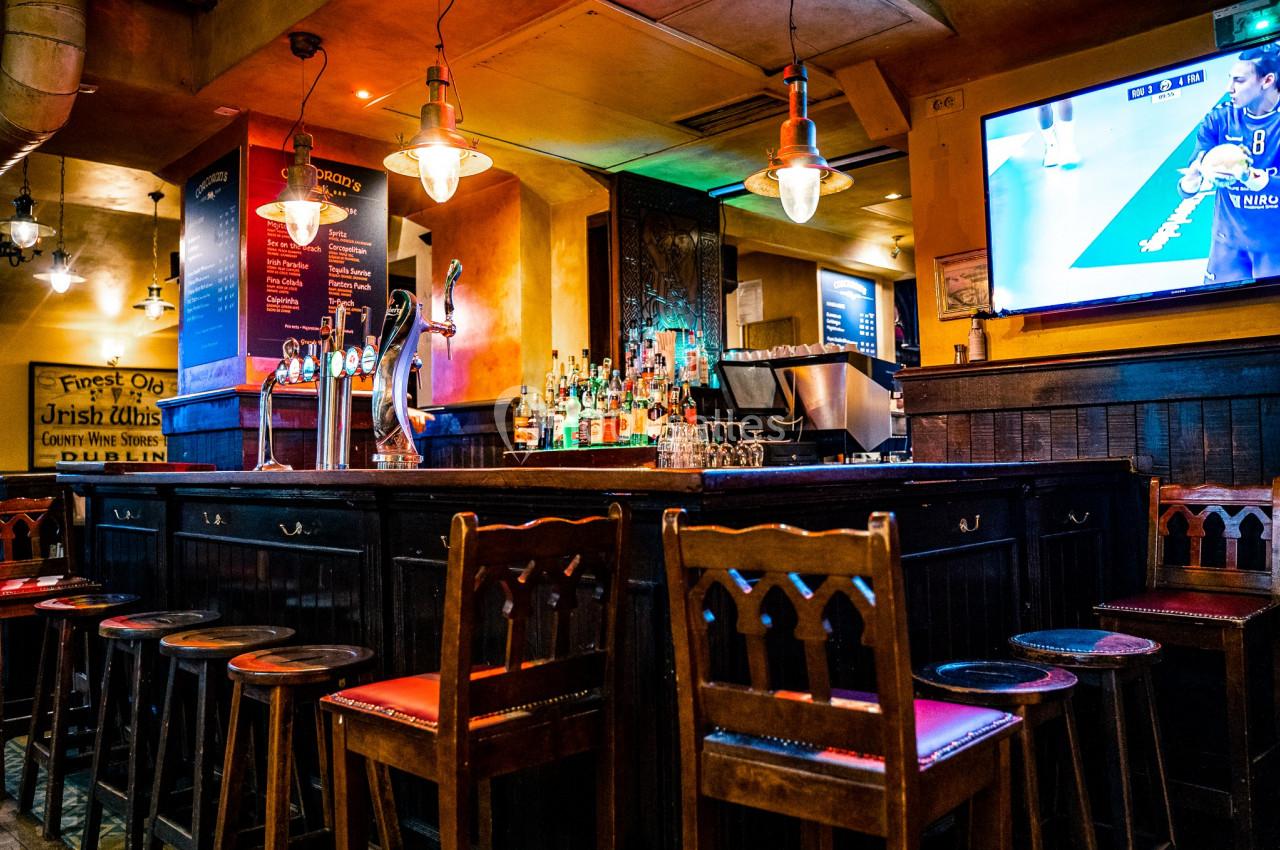 Intérieur d'un pub avec un bar en bois, des tabourets, des lumières suspendues et un écran diffusant un match.