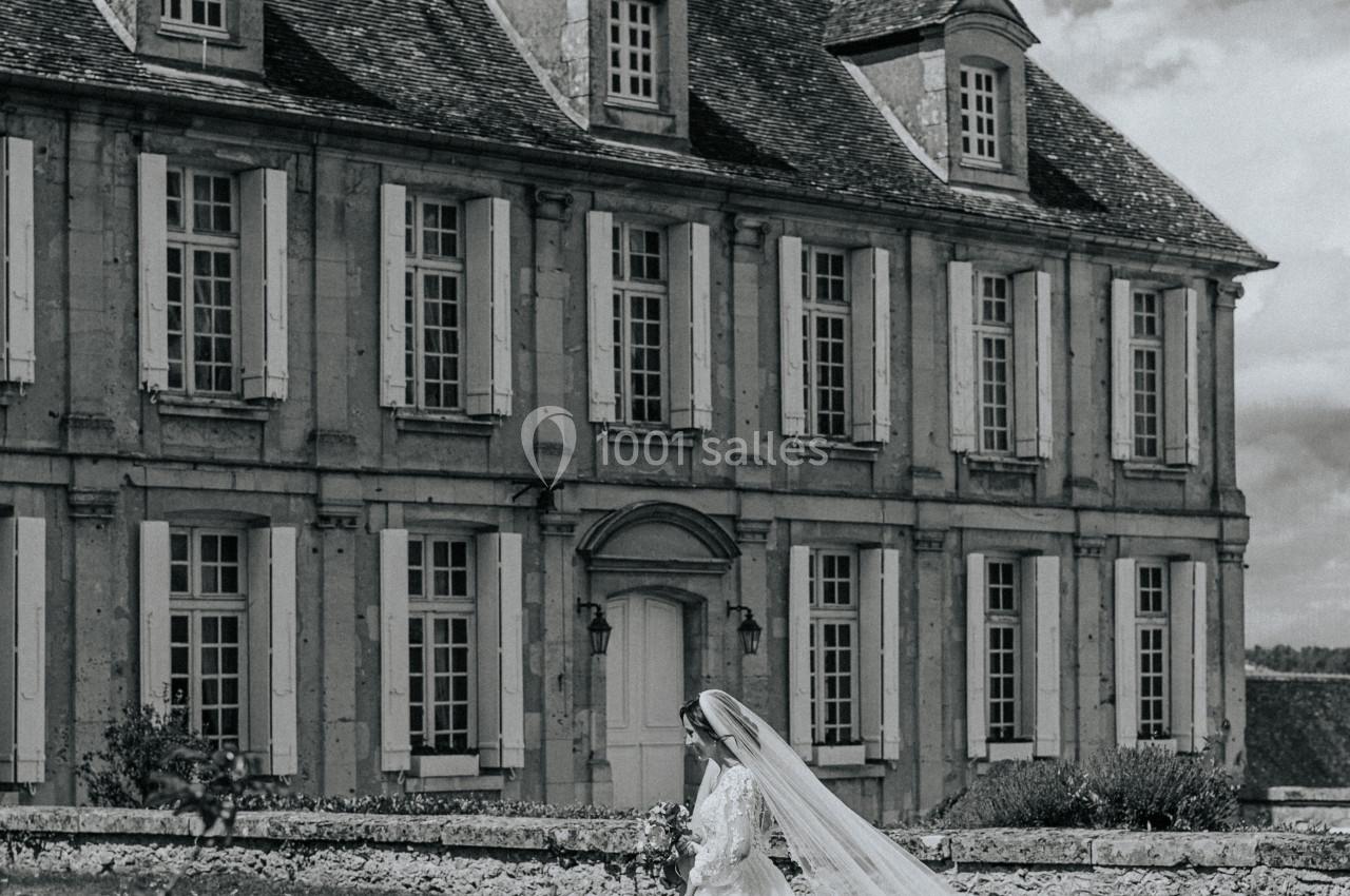 Une mariée en robe blanche marche sur une pelouse devant un grand bâtiment ancien en pierre.