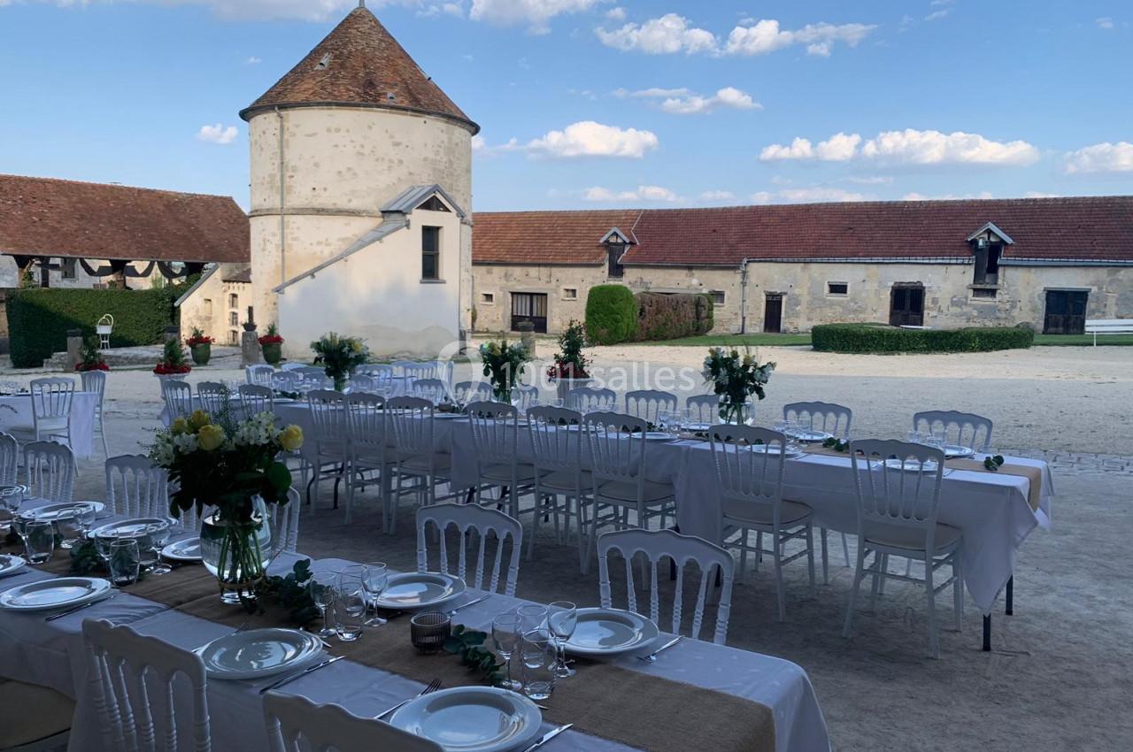 Tables dressées avec nappes blanches et décorations florales dans une cour de ferme avec bâtiments en pierre et ciel dégagé.