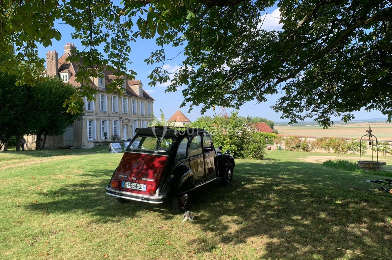 Une voiture ancienne garée sur une pelouse ombragée devant une maison de campagne avec vue sur un paysage rural.