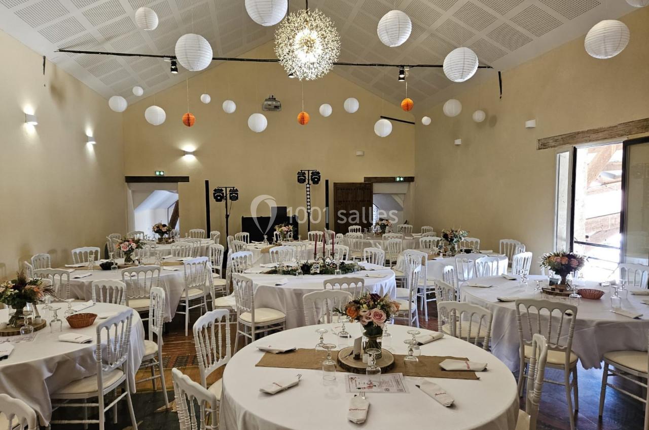 Salle décorée pour un événement avec tables rondes dressées, chaises blanches et lanternes suspendues au plafond.