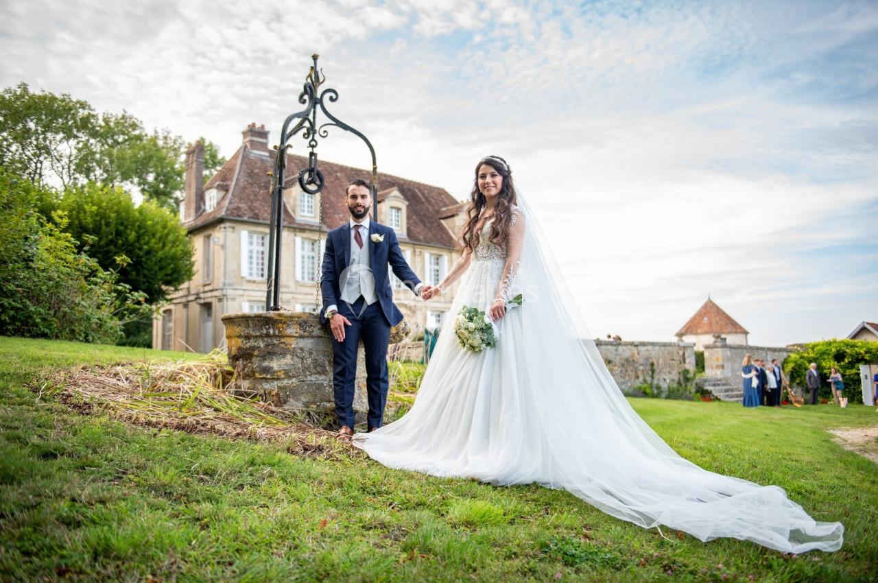Un couple de mariés pose près d'un puits dans un jardin, avec une maison ancienne en arrière-plan.