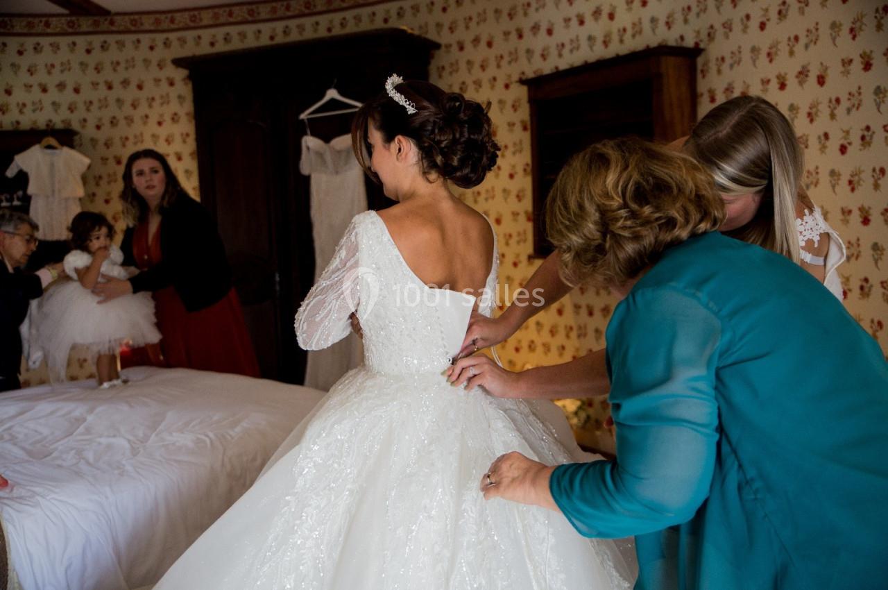 Une mariée en robe blanche est aidée par deux femmes pour ajuster sa tenue dans une chambre décorée de papier peint fleuri.