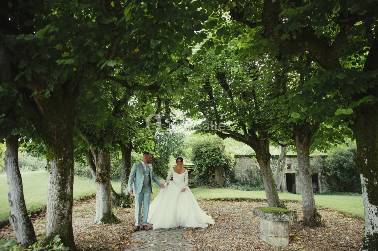Un couple en tenue de mariage pose sous des arbres dans un jardin verdoyant avec un banc en pierre à proximité.
