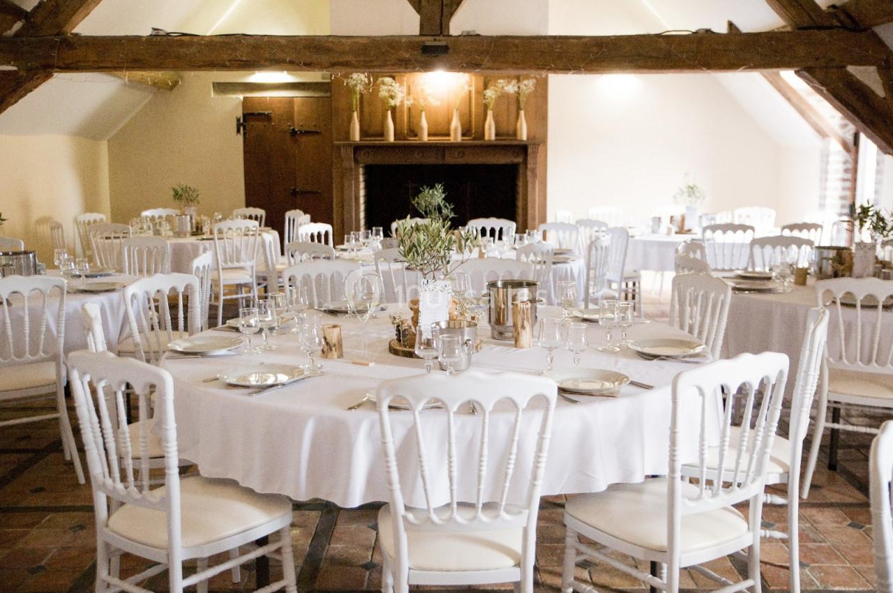Salle de réception lumineuse avec tables rondes dressées, nappes blanches et chaises assorties sous une charpente en bois.