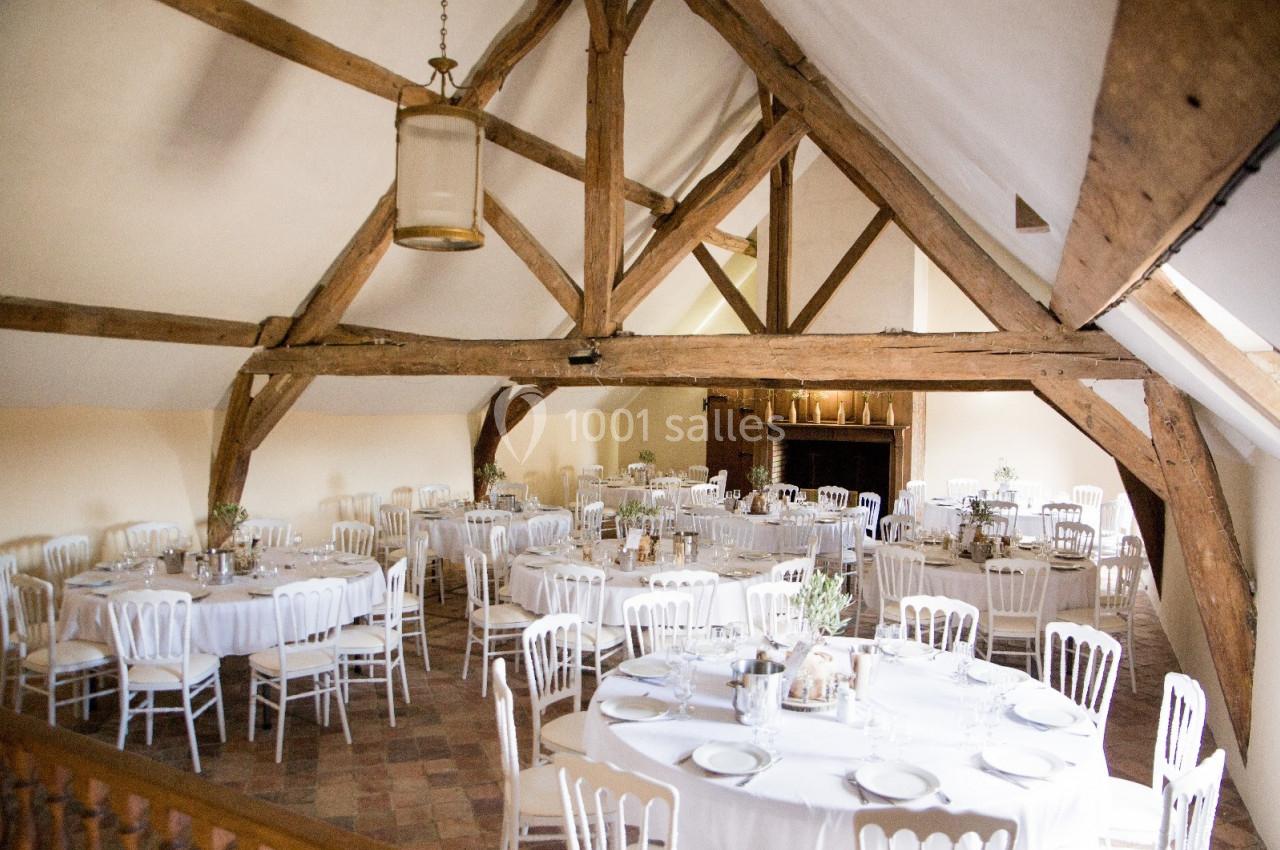 Salle de réception lumineuse avec poutres apparentes, tables rondes dressées et chaises blanches alignées.