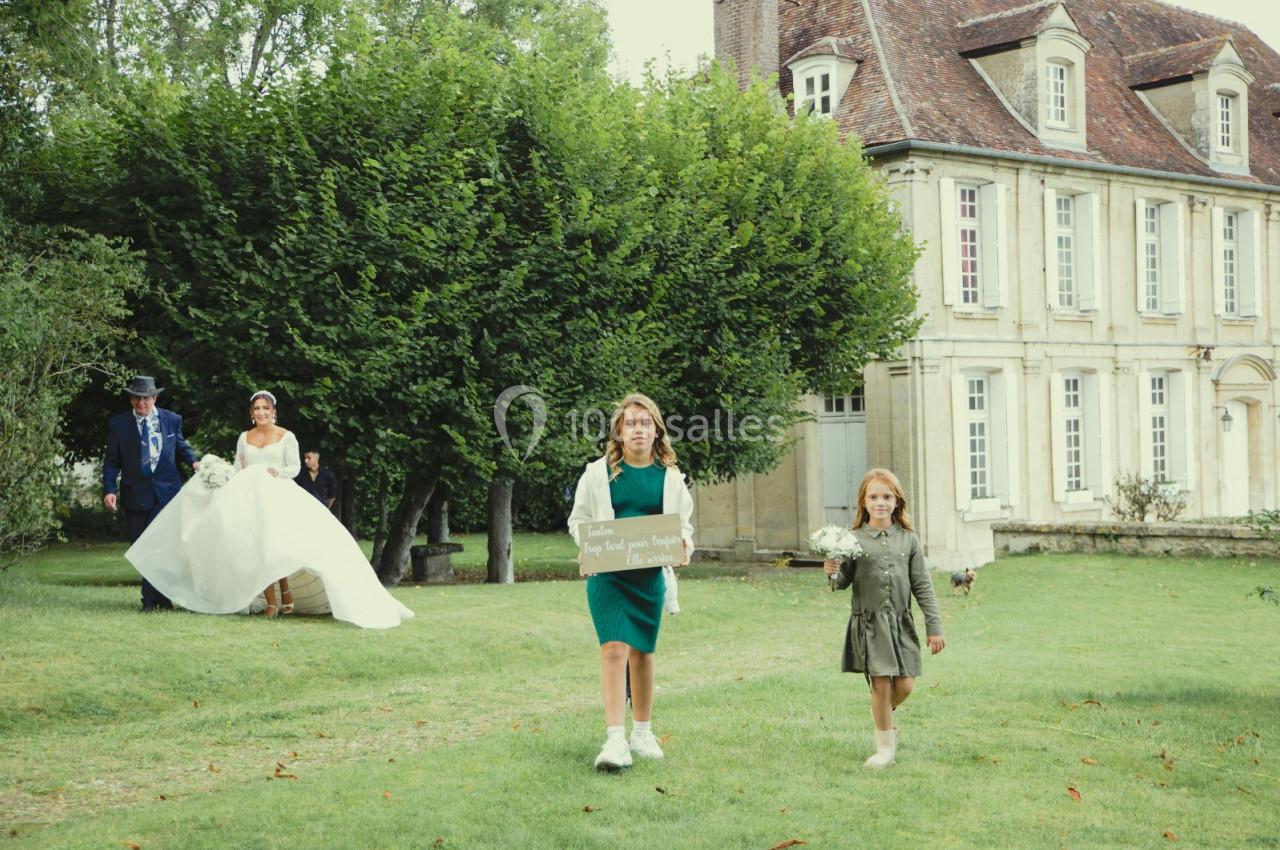 Deux enfants marchent sur une pelouse devant une mariée et un homme, près d'une grande maison ancienne.