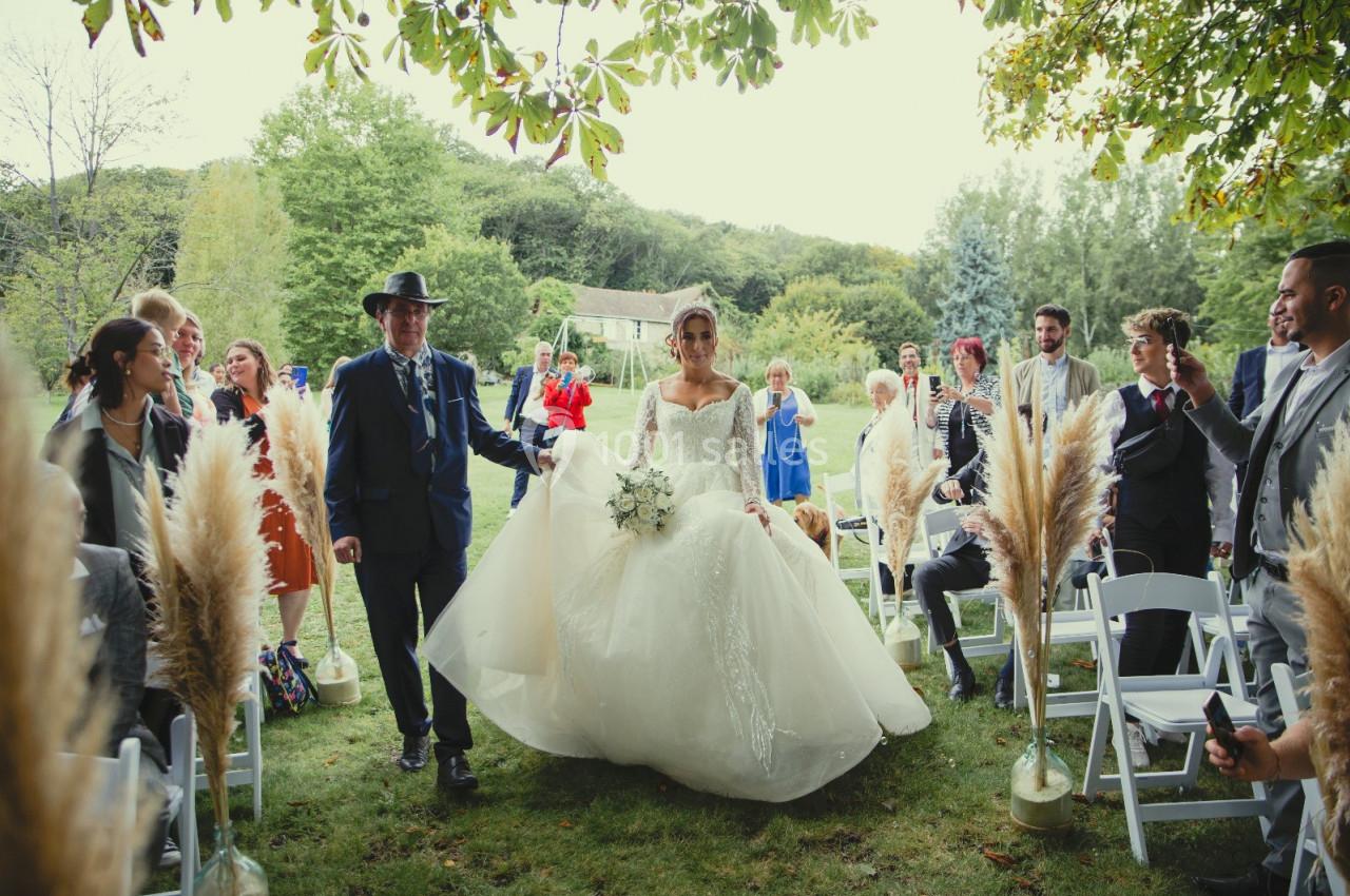 Une mariée en robe blanche avance dans une allée extérieure bordée d'invités assis sous des arbres.