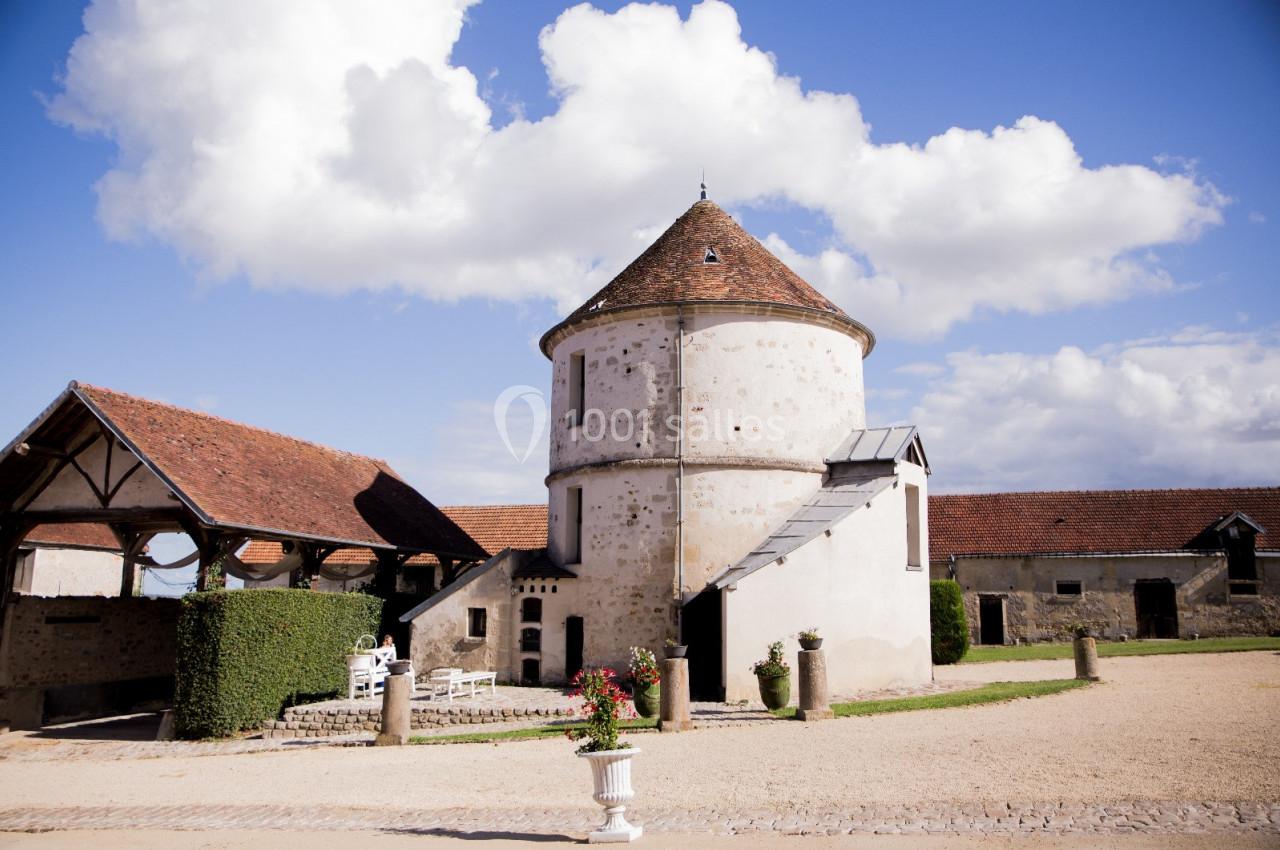 Bâtiment en pierre avec toit en tuiles, entouré de dépendances et d'une cour gravillonnée sous un ciel bleu.