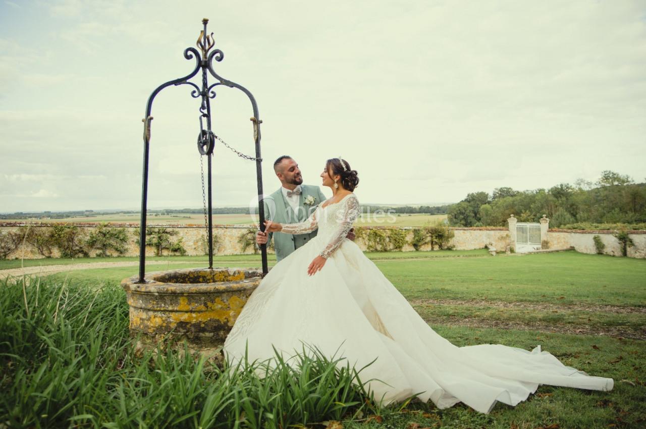 Un couple en tenue de mariage se tient près d'un puits ancien dans un jardin verdoyant.