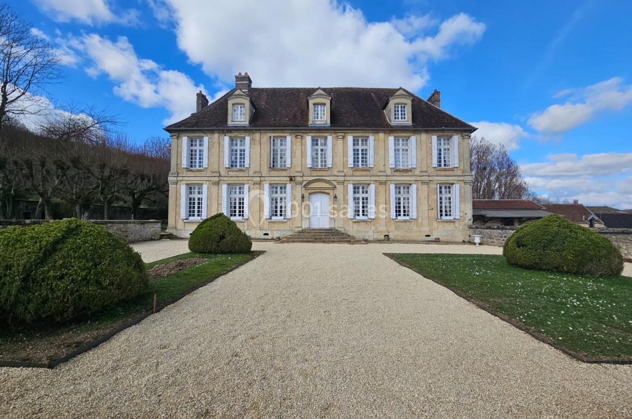 Façade d'un manoir en pierre avec toit en ardoise, entouré d'un jardin et d'une allée gravillonnée sous un ciel bleu.