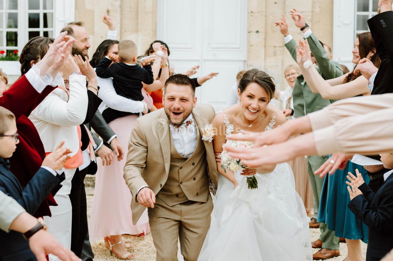 Un couple de mariés souriants sort entouré d'invités applaudissant devant un bâtiment ancien.