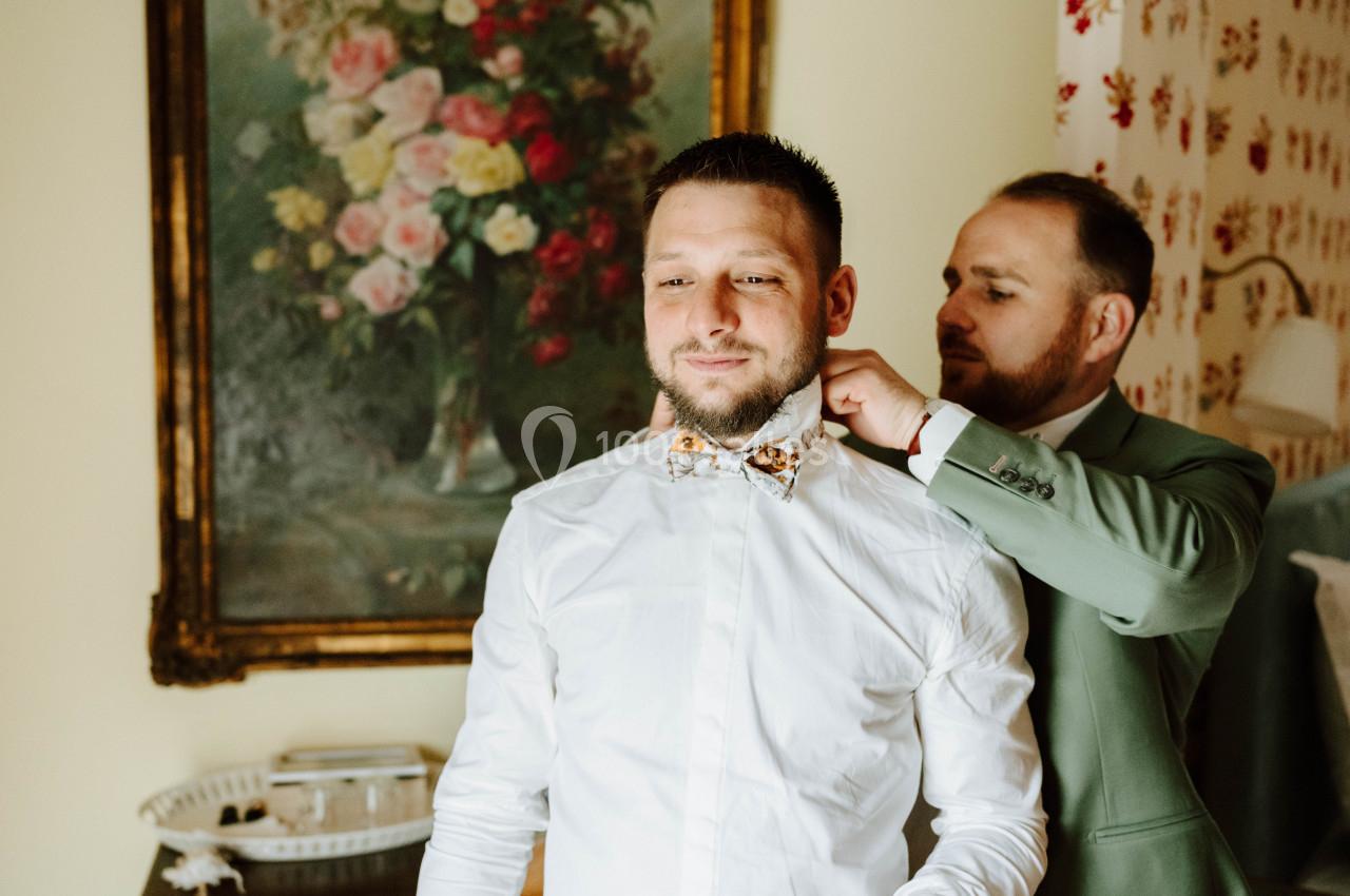 Un homme ajuste le nœud papillon d'un autre homme dans une pièce décorée avec un tableau floral et des motifs rouges.
