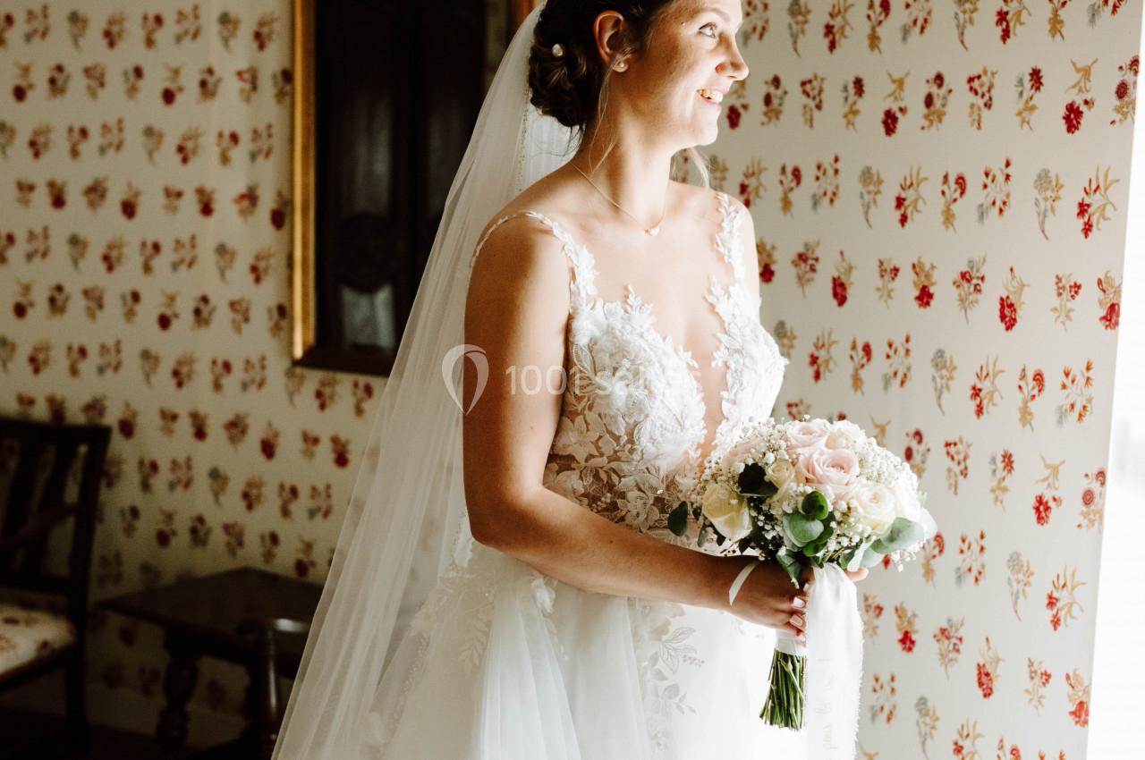 Une mariée en robe blanche tient un bouquet de fleurs, debout près d'une fenêtre dans une pièce au papier peint fleuri.