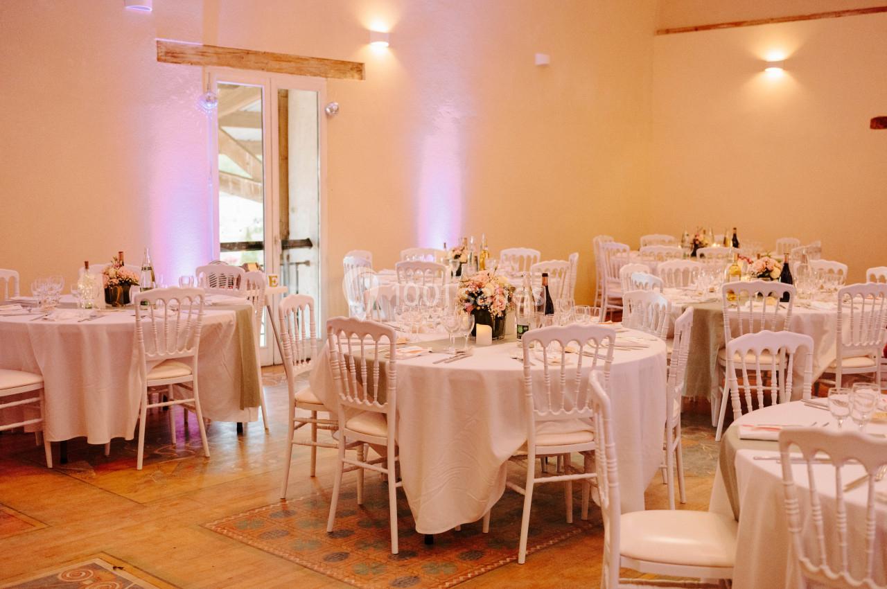 Salle de réception décorée avec des tables rondes, nappes blanches, chaises élégantes et arrangements floraux.