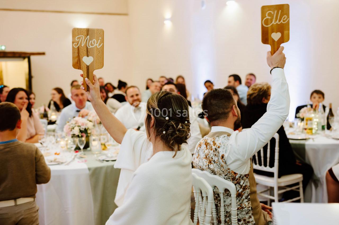 Un couple assis de dos lors d'un mariage lève des pancartes en bois indiquant ’Moi’ et ’Elle’ devant des invités.